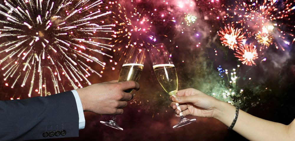 Two hands toasting with champagne glasses against a vibrant backdrop of colorful fireworks, creating a festive and celebratory atmosphere.