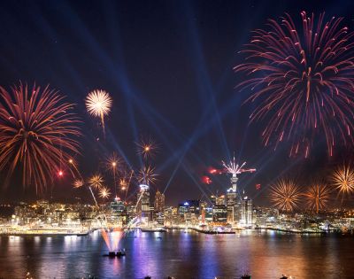 Nighttime cityscape with vibrant red and gold fireworks above a harbor, reflecting in water. Skyline illuminated; boats visible. Celebratory and festive atmosphere.