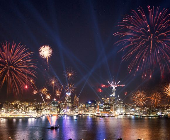 Nighttime cityscape with vibrant red and gold fireworks above a harbor, reflecting in water. Skyline illuminated; boats visible. Celebratory and festive atmosphere.