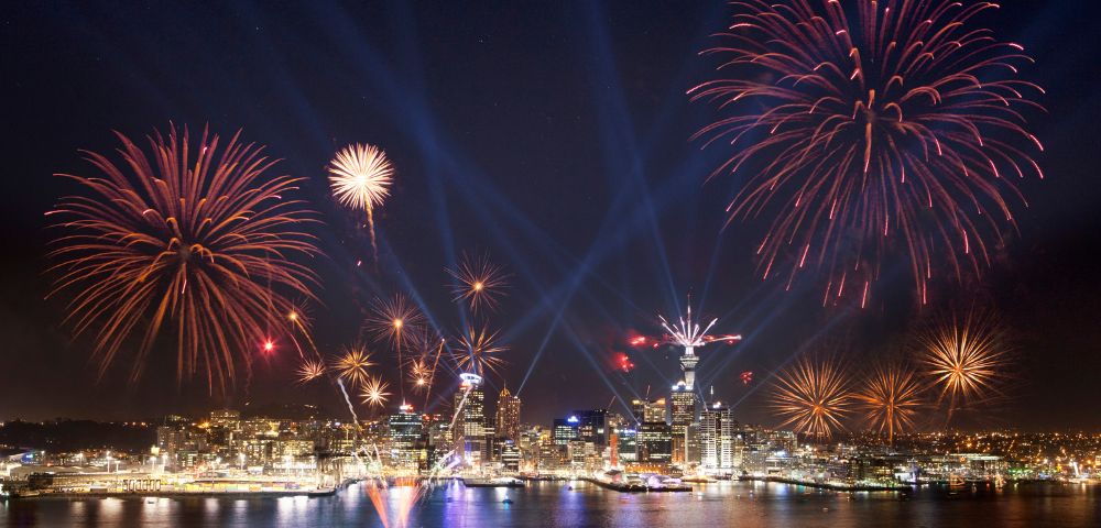 Nighttime cityscape with vibrant red and gold fireworks above a harbor, reflecting in water. Skyline illuminated; boats visible. Celebratory and festive atmosphere.