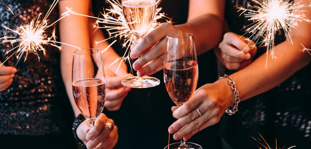 Hands holding sparklers and champagne glasses, creating a festive, celebratory atmosphere. The scene is lively with sparkling lights and bubbly drinks.