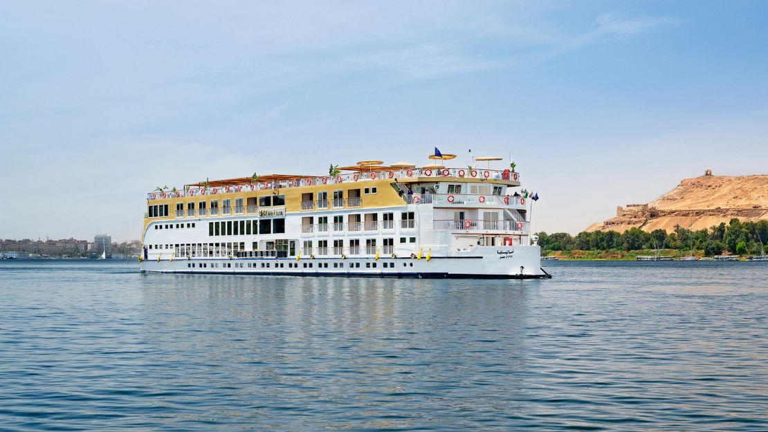 A Nile River cruise ship glides smoothly on the water, surrounded by lush greenery and a clear blue sky.