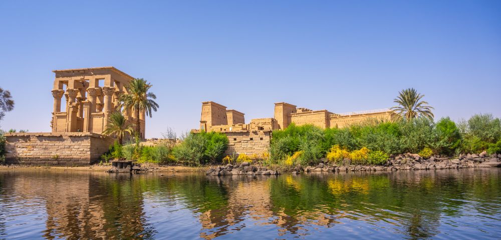 Ancient Egyptian temple ruins beside lush greenery and palm trees reflecting in calm waters under a clear blue sky.