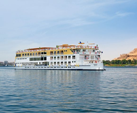 A Nile River cruise ship glides smoothly on the water, surrounded by lush greenery and a clear blue sky.