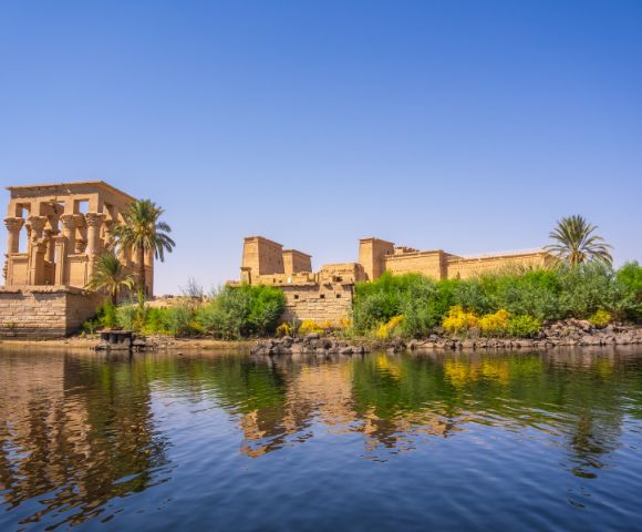 Ancient Egyptian temple ruins beside lush greenery and palm trees reflecting in calm waters under a clear blue sky.