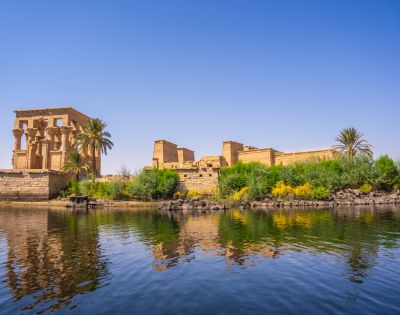 Ancient Egyptian temple ruins beside lush greenery and palm trees reflecting in calm waters under a clear blue sky.
