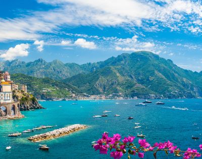 Scenic coastal town with colorful cliffside buildings overlooking a vibrant blue sea, dotted with boats and yachts. Lush green mountains rise in the background, and bright pink flowers bloom in the foreground.