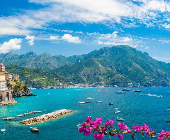 Scenic coastal town with colorful cliffside buildings overlooking a vibrant blue sea, dotted with boats and yachts. Lush green mountains rise in the background, and bright pink flowers bloom in the foreground.