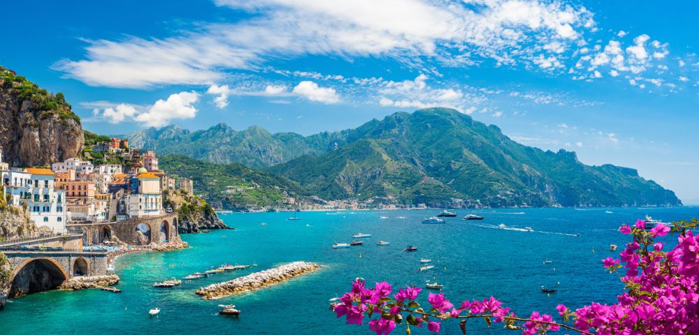 Scenic coastal town with colorful cliffside buildings overlooking a vibrant blue sea, dotted with boats and yachts. Lush green mountains rise in the background, and bright pink flowers bloom in the foreground.