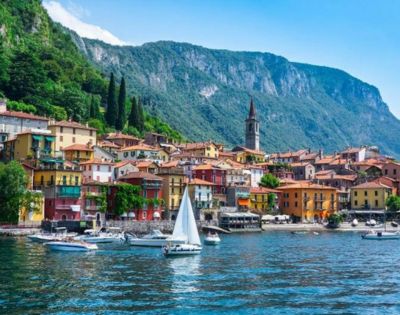Coastal town with colorful buildings, a church tower, and lush green hills behind. Boats and a sailboat are on a calm blue lake, invoking serenity.