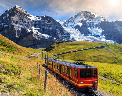 A red and yellow train travels through lush green hills with majestic snow-capped mountains in the background under a clear blue sky.