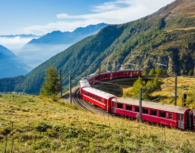 Red train winding through lush, mountainous landscape on a sunny day, with blue sky and distant hills creating a serene and picturesque scene.
