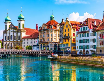Colorful historic buildings line the vibrant lakefront in Lucerne, Switzerland, reflecting in the clear water. The scene is lively and picturesque.