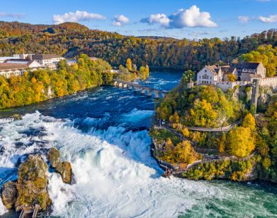 Aerial view of Rhine Falls in Switzerland, surrounded by autumn trees. A historic building sits on a cliff, overlooking the cascading water. Bright, scenic.