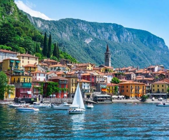 Coastal town with colorful buildings, a church tower, and lush green hills behind. Boats and a sailboat are on a calm blue lake, invoking serenity.