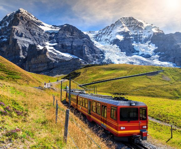 A red and yellow train travels through lush green hills with majestic snow-capped mountains in the background under a clear blue sky.