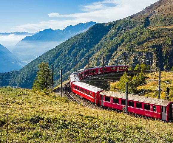 Red train winding through lush, mountainous landscape on a sunny day, with blue sky and distant hills creating a serene and picturesque scene.