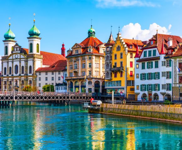 Colorful historic buildings line the vibrant lakefront in Lucerne, Switzerland, reflecting in the clear water. The scene is lively and picturesque.