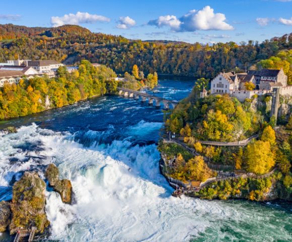 Aerial view of Rhine Falls in Switzerland, surrounded by autumn trees. A historic building sits on a cliff, overlooking the cascading water. Bright, scenic.