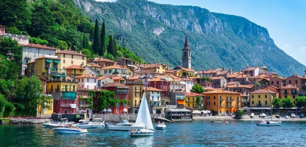 Coastal town with colorful buildings, a church tower, and lush green hills behind. Boats and a sailboat are on a calm blue lake, invoking serenity.