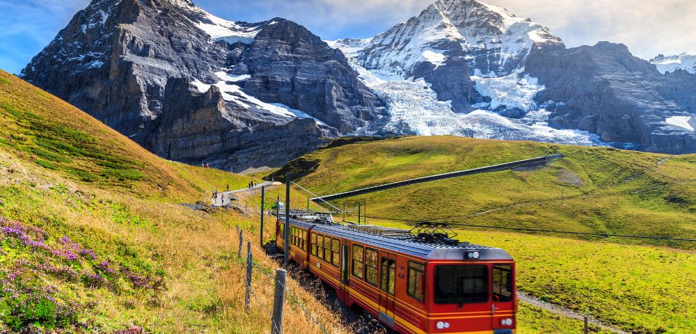A red and yellow train travels through lush green hills with majestic snow-capped mountains in the background under a clear blue sky.