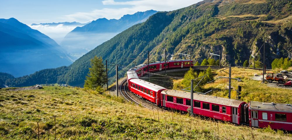Red train winding through lush, mountainous landscape on a sunny day, with blue sky and distant hills creating a serene and picturesque scene.