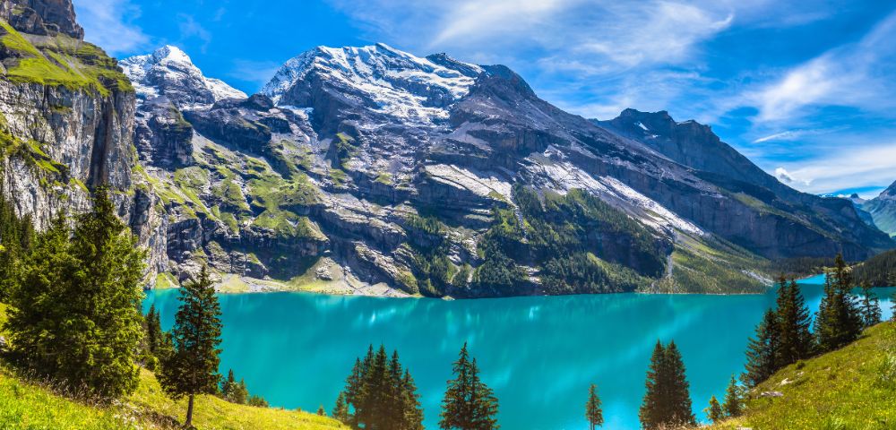 A serene mountain landscape with a clear turquoise lake bordered by lush green pine trees. Snow-capped peaks under a bright blue sky enhance the scene's tranquility.