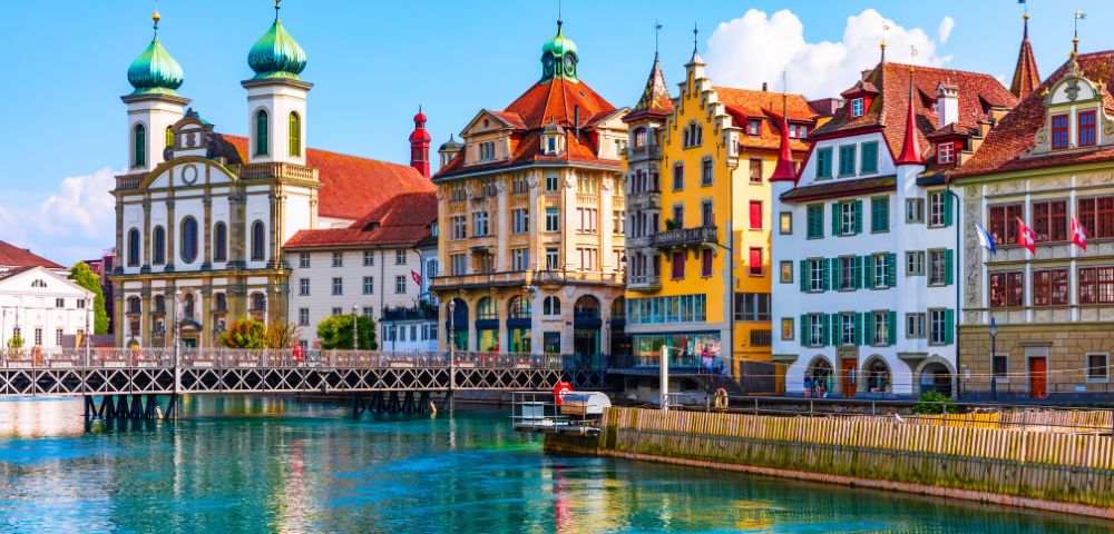 Colorful historic buildings line the vibrant lakefront in Lucerne, Switzerland, reflecting in the clear water. The scene is lively and picturesque.