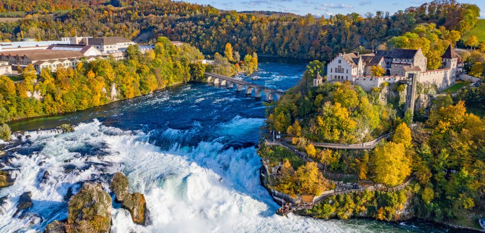 Aerial view of Rhine Falls in Switzerland, surrounded by autumn trees. A historic building sits on a cliff, overlooking the cascading water. Bright, scenic.