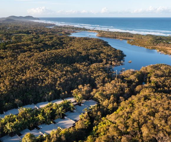 Aerial view of a coastline with a river snaking through a forest, a sandy beach, and the ocean, with a resort visible in the foreground.