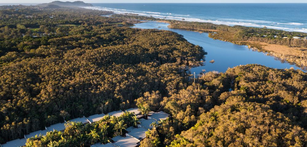 Aerial view of a coastline with a river snaking through a forest, a sandy beach, and the ocean, with a resort visible in the foreground.