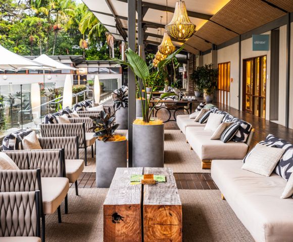 An upscale outdoor lounge area with neutral colored couches and patterned throw pillows lining a wood floor, accented by a wooden coffee table with plants in concrete pots and overhead light fixtures, with views of lush greenery and a small outdoor dining area in the background.