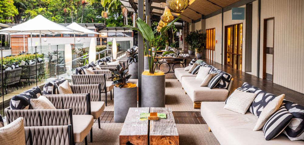 An upscale outdoor lounge area with neutral colored couches and patterned throw pillows lining a wood floor, accented by a wooden coffee table with plants in concrete pots and overhead light fixtures, with views of lush greenery and a small outdoor dining area in the background.