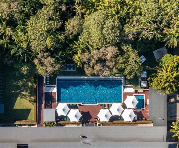 Aerial view of a lush resort pool surrounded by greenery, featuring lounge chairs and shaded cabanas on a sunny day.