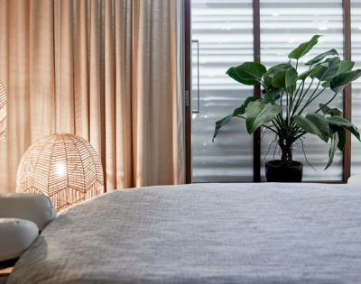Cozy bedroom corner featuring soft curtains, rattan lamps, a plant, and a neatly made bed with a textured gray cover.