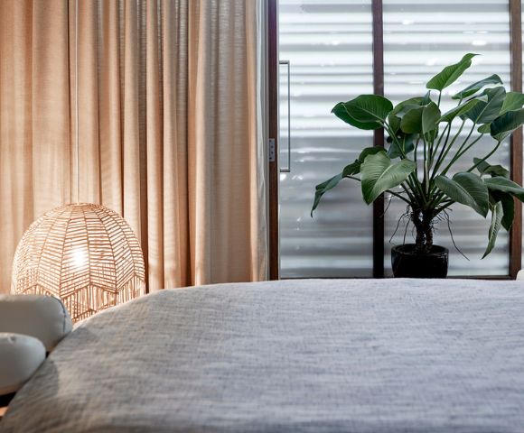Cozy bedroom corner featuring soft curtains, rattan lamps, a plant, and a neatly made bed with a textured gray cover.