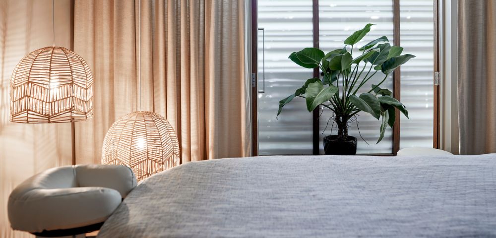 Cozy bedroom corner featuring soft curtains, rattan lamps, a plant, and a neatly made bed with a textured gray cover.
