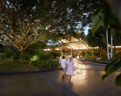 A couple strolls towards a warmly lit entrance, under a large tree adorned with lights, surrounded by lush greenery in the evening.
