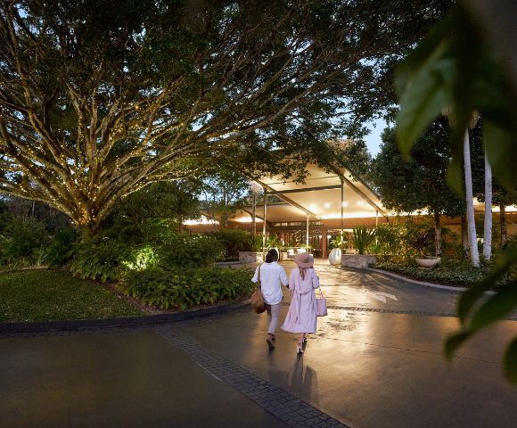 A couple strolls towards a warmly lit entrance, under a large tree adorned with lights, surrounded by lush greenery in the evening.