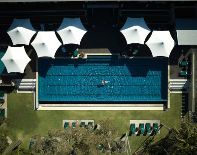 Aerial view of a serene swimming pool with a swimmer, surrounded by sunshades and lounge chairs on a sunny day.