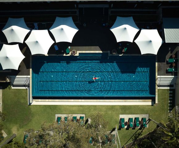 Aerial view of a serene swimming pool with a swimmer, surrounded by sunshades and lounge chairs on a sunny day.