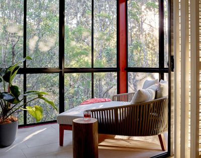 Sunlit interior with outdoor view, featuring a modern lounge chair with cushions, a small wooden side table with a drink, a potted plant, and a large window overlooking a forest.