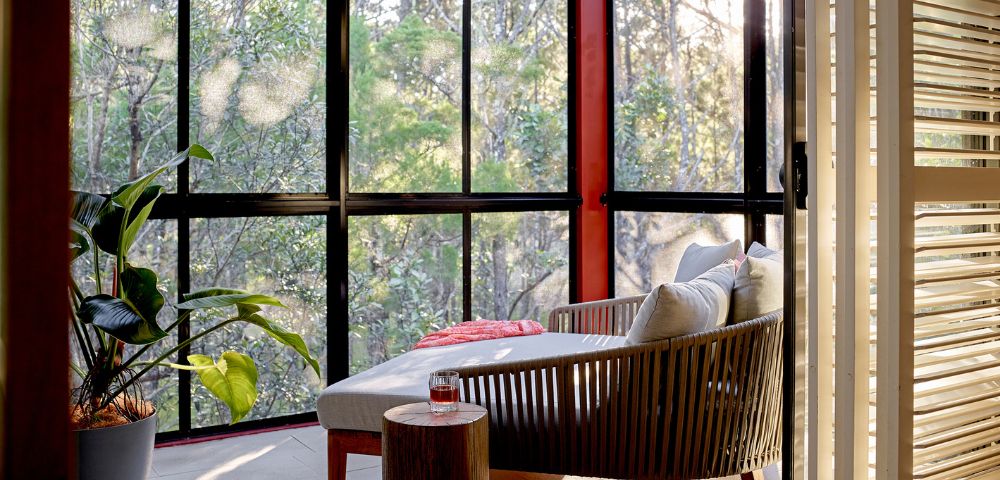 Sunlit interior with outdoor view, featuring a modern lounge chair with cushions, a small wooden side table with a drink, a potted plant, and a large window overlooking a forest.