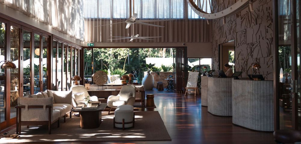 Elegant hotel lobby with open doors leading to a garden, featuring a reception desk, seating area with sofas and armchairs, and decorative lighting.