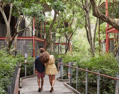 A couple walks arm in arm down a wooden boardwalk flanked by lush greenery, passing a red metal framed building with balconies and lush green foliage growing between buildings.