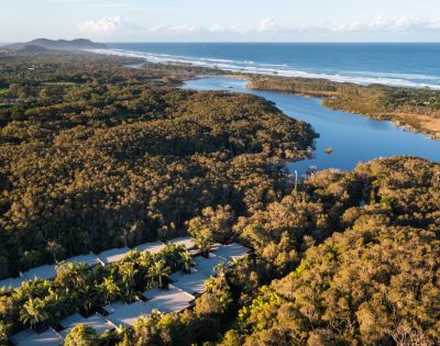 Aerial view of a coastline with a river snaking through a forest, a sandy beach, and the ocean, with a resort visible in the foreground.