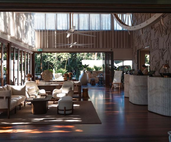 Elegant hotel lobby with open doors leading to a garden, featuring a reception desk, seating area with sofas and armchairs, and decorative lighting.
