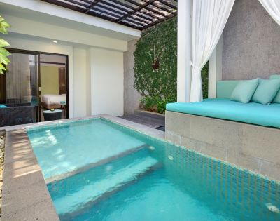 Outdoor luxury resort pool with turquoise water, built-in lounge seating with aqua cushions, a canopy of sheer white curtains, and a background of climbing ivy and a partially visible bedroom.