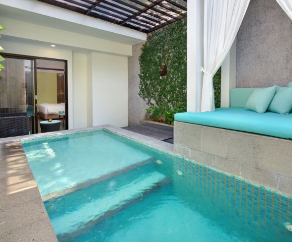 Outdoor luxury resort pool with turquoise water, built-in lounge seating with aqua cushions, a canopy of sheer white curtains, and a background of climbing ivy and a partially visible bedroom.