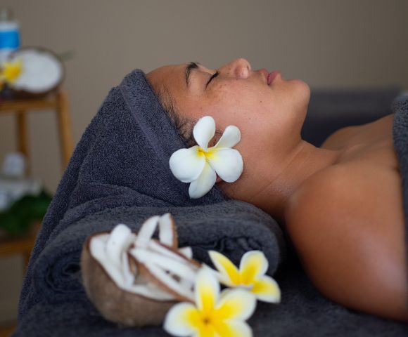A captivating image displays a tranquil scene, where a woman lies on a dark blue towel, eyes closed, surrounded by white flowers. The images elegance shines through the blurred spa background, evoking a sense of relaxation and self-care.